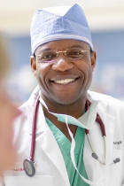 Close-up picture of world-renowned heart surgeon Dr. Nche Zama. He is wearing a blue surgeons cap, white doctor's robe over green scrubs, a facial mask hanging around his neck and a stethoscope