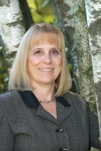 Portrait head shot of Bonnie Schwartz, a woman with blonde shoulder length hair, wearing a grey and black tweed blazer