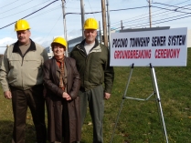 Three individuals wear yellow hard hats in front of the ground breaking site for the Pocono Township Sewer Project