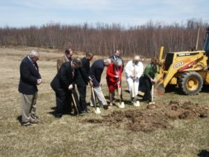 A group of individuals are in a field with shovels digging into the ground with a yellow construction vehicle in the background.