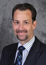 Head shot of Michael Baxter. He has short dark hair and a goatee facial hair. He's wearing a striped button up shirt with a blue tie, and a pinstripe grey blazer.