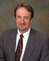 Head shot of Paul Canevari. He has short dark hair and is wearing a white button up dress shirt with a red patterned tie and a grey blazer on top.
